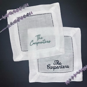 May include: Two white linen coasters with embroidered text that reads "The Carpenters". The coasters have a square shape with a decorative stitched border. One coaster has green text, and the other has black text. Lavender sprigs are placed around the coasters.