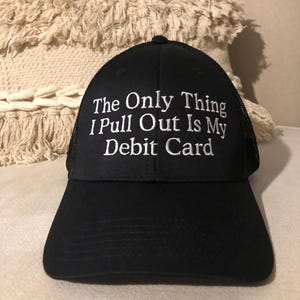 May include: Black trucker hat with the text "The Only Thing I Pull Out Is My Debit Card" embroidered in white. The hat has a curved brim and a mesh back.