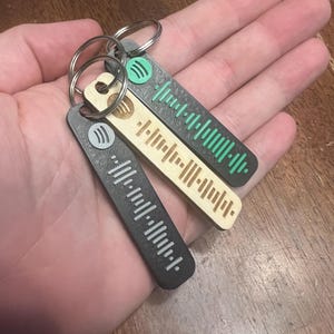 May include: Three keychains with Spotify code designs. One is black with white lines, another is light wood with brown lines, and the third is black with green lines. Each keychain has a silver keyring and the Spotify logo.