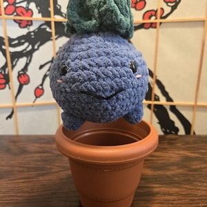 Puede incluir: Una criatura azul hecha a mano con ganchillo con una parte superior verde, una cara sonriente y pequeños ojos negros. El peluche se sostiene sobre una maceta de plástico de color terracota, lo que sugiere un tema de plantas.