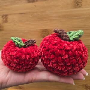 May include: Two red crocheted apples with green leaves and brown stems. The apples are made of a soft, textured yarn. One apple is smaller than the other. The apples are held in a hand against a wooden background.