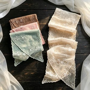 May include: A collection of rectangular lace scarves featuring intricate white floral embroidery and scalloped edges. The set includes folded scarves in shades of brown, pink, sage green, and cream, each displaying a delicate mesh texture and floral patterns.