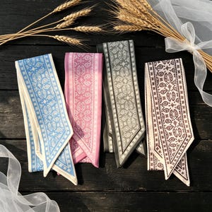 Könnte beinhalten: Vier gemusterte Seidenschals in Blau-, Rosa-, Grau- und Brauntönen. Jeder Schal hat ein Paisley-Design und einen kontrastierenden Rand. Die Schals sind auf einer dunklen Holzoberfläche angeordnet, mit Weizenähren und Schleife im Hintergrund.