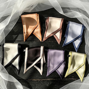 May include: Seven satin neck scarves arranged in a grid, each featuring a solid colour with a contrasting border. The colours include tan, cream, light blue, white, black, lavender, and pale yellow. Each scarf has a pointed, folded design.