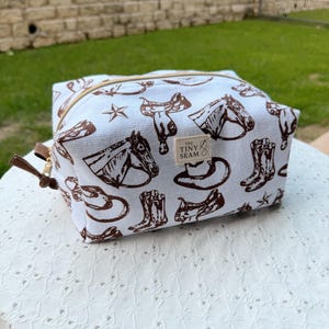 May include: A white canvas cosmetic bag with a brown cowboy-themed pattern. The pattern includes horses, saddles, boots, and stars. The bag has a gold zipper and a small tag that reads "The Tiny Seam."