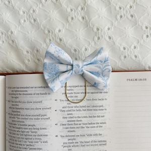 May include: A light blue and white floral patterned bow tie with a gold-colored paper clip, resting on an open book. The book's pages display printed text from Psalm 18:50.