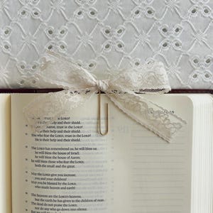 May include: A cream lace bow bookmark with a gold paperclip, placed on the open pages of a book. The book's pages show printed text. The background features white eyelet fabric.