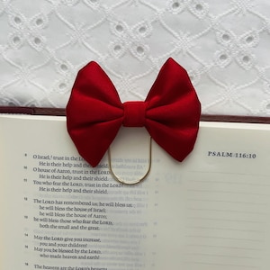 May include: A bright red bow bookmark with a gold-coloured clip, placed on the open pages of a book. The text on the page reads "PSALM 116:10". The background is a white, patterned fabric.