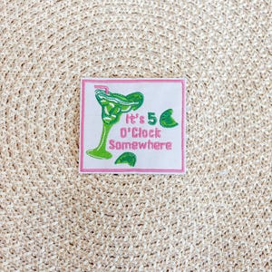May include: Square patch with a pink border and the text "It's 5 O'Clock Somewhere." The patch features a green margarita glass and lime slices. The patch is on a beige woven surface.