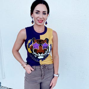 May include: A sleeveless knit top featuring a tiger face design. The top is split vertically, with a navy blue side and a yellow side. The tiger face has purple star-shaped sunglasses. The person is wearing brown trousers and a white watch.