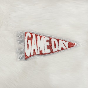 Könnte beinhalten: Ein rot-weißes Wimpelbanner mit den Worten "GAME DAY" in weißen Blockbuchstaben. Der Wimpel ist silberfarben umrandet und hat eine silberne Fransen. Die Flagge befindet sich in einer transparenten Plastiktüte.