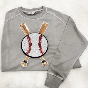 May include: Grey sweatshirt with a baseball motif. The baseball is white with red stitching, crossed with gold baseball bats. The design is embellished with sequins. The sweatshirt has a crew neck and ribbed cuffs.