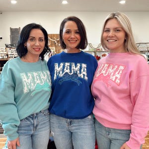 May include: Three women wearing sweatshirts with the word "MAMA" in white block letters. The sweatshirts are in light blue, royal blue, and pink. Each sweatshirt has a bow graphic. The women are wearing blue jeans.