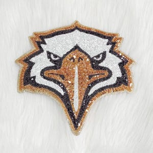 May include: A sequined eagle head patch in gold, white, and black. The patch is shaped like a shield and features a detailed design of an eagle's face. The sequins create a textured, shimmering effect, perfect for embellishing apparel or accessories.