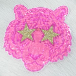 May include: A pink sequined tiger face patch with gold glitter star-shaped sunglasses. The patch is detailed with pink sequins and has a white backing. The tiger's face is centered, with the star-shaped sunglasses covering the eyes.