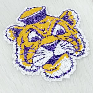 May include: A sequined patch in the shape of a tiger's head, featuring yellow and purple sequins. The tiger is wearing a purple hat. The patch has a white border.