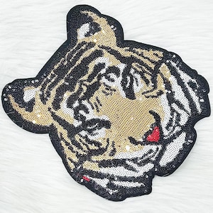 Può includere: Una toppa di paillettes a forma di testa di tigre. La toppa presenta un bordo nero, paillettes dorate e nere per il viso e le strisce della tigre e paillettes bianche per le guance. La bocca della tigre è rossa.