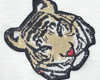 May include: A sequined patch in the shape of a tiger's head. The patch features a black border, gold and black sequins for the tiger's face and stripes, and white sequins for the tiger's cheeks. The tiger's mouth is red.