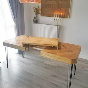 Op de afbeelding: Een licht houten bureau met een uniek, gebogen ontwerp. Het bureau heeft een visgraatpatroon op het tafelblad en wordt ondersteund door zwarte haarspeldpoten. Het bureau heeft een verhoogd gedeelte in het midden en staat in een kamer met een lichtgrijze vloer.