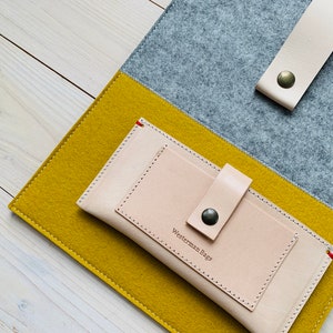 May include: A light beige leather wallet with a flap closure and a snap button. The wallet has a pocket on the front and the text "Westerman Bags" is embossed on the front. The wallet is sitting on top of a yellow and gray felt fabric.