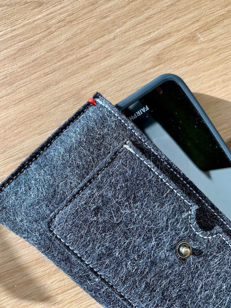 May include: A black felt phone case with a white stitch detail and a small metal button. The case has a pocket on the front and the text "FAIRPH" is visible on the phone.