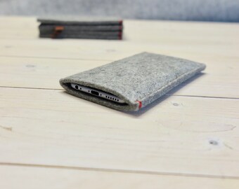 May include: A gray felt phone case with red stitching. The case is lying on a white wooden surface.