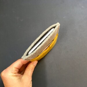 May include: A grey felt phone case with a yellow felt lining. The case is folded in half and the phone is inside.