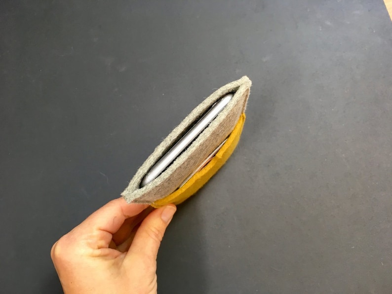 May include: A grey felt phone case with a yellow felt lining. The case is folded in half and the phone is inside.