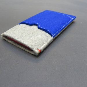 May include: A small grey felt wallet with a blue felt flap. The wallet has white stitching around the edges and a red accent on the flap.