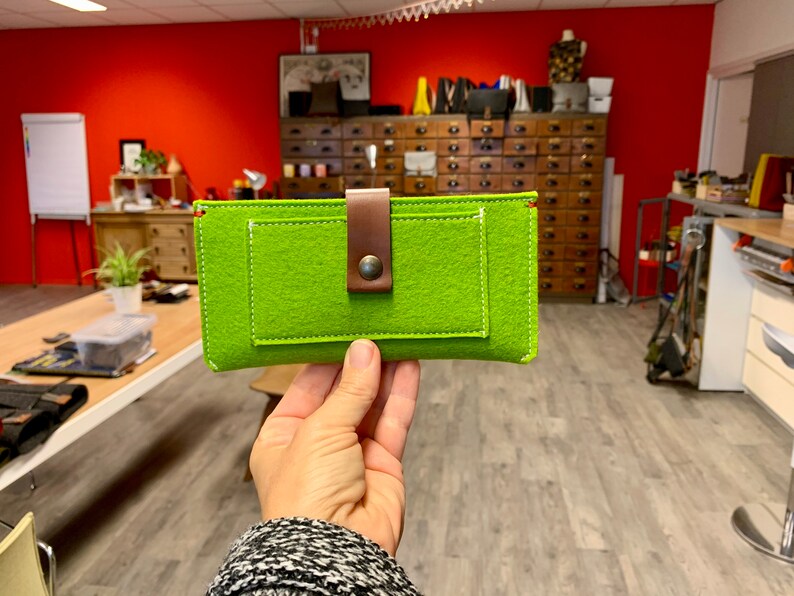 May include: A bright green felt phone case with a brown leather strap and a metal button. The case has a small pocket on the front.