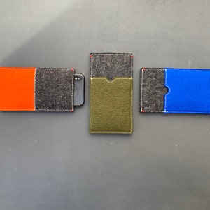 May include: Three felt phone cases, each with a different coloured insert. The first case has a black felt exterior with an orange felt insert. The second case has a black felt exterior with a green felt insert. The third case has a black felt exterior with a blue felt insert.