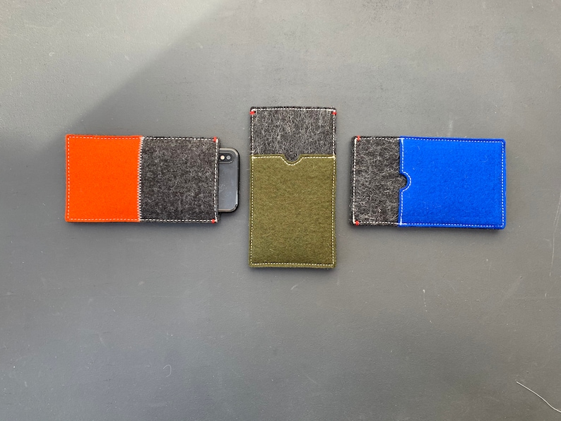 May include: Three felt phone cases, each with a different coloured insert. The first case has a black felt exterior with an orange felt insert. The second case has a black felt exterior with a green felt insert. The third case has a black felt exterior with a blue felt insert.