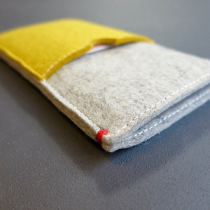 May include: A grey felt phone case with a yellow felt flap. The case has white stitching around the edges and a red stitch detail at the corner.