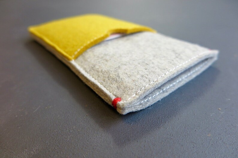 May include: A grey felt phone case with a yellow felt flap. The case has white stitching around the edges and a red stitch detail at the corner.