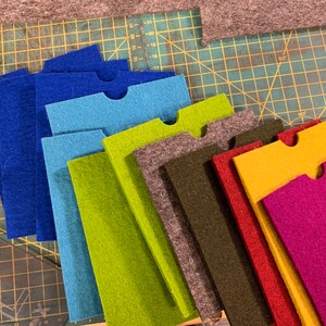 May include: A set of ten felt fabric pieces in various colours, including blue, green, brown, red, yellow, and purple. Each piece has a cut-out shape in the top centre.
