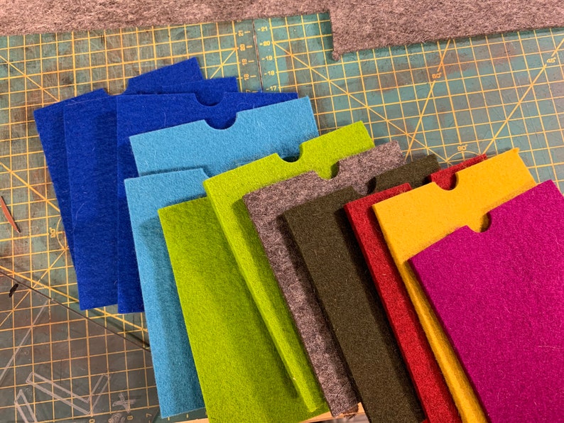 May include: A set of ten felt fabric pieces in various colours, including blue, green, brown, red, yellow, and purple. Each piece has a cut-out shape in the top centre.