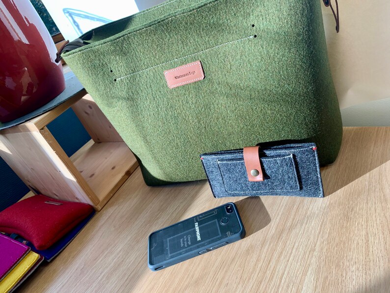 May include: A green felt tote bag with a brown leather handle and a label that says "Wanderlust Co.". A gray felt wallet with a brown leather strap and a black felt phone case are also pictured.