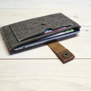 May include: A gray felt phone case with a brown leather strap and a snap closure. The case has a pocket for cards and a phone is visible inside.