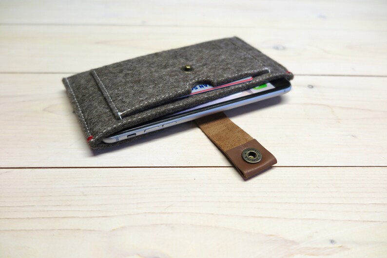 May include: A gray felt phone case with a brown leather strap and a snap closure. The case has a pocket for cards and a phone is visible inside.