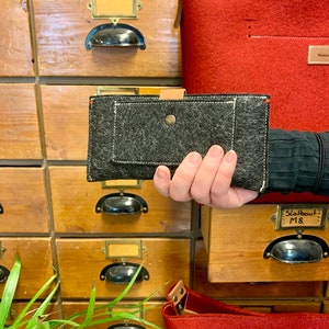 May include: A black felt wallet with a brown leather flap and a white stitch detail. The wallet is held in a hand against a background of wooden drawers.
