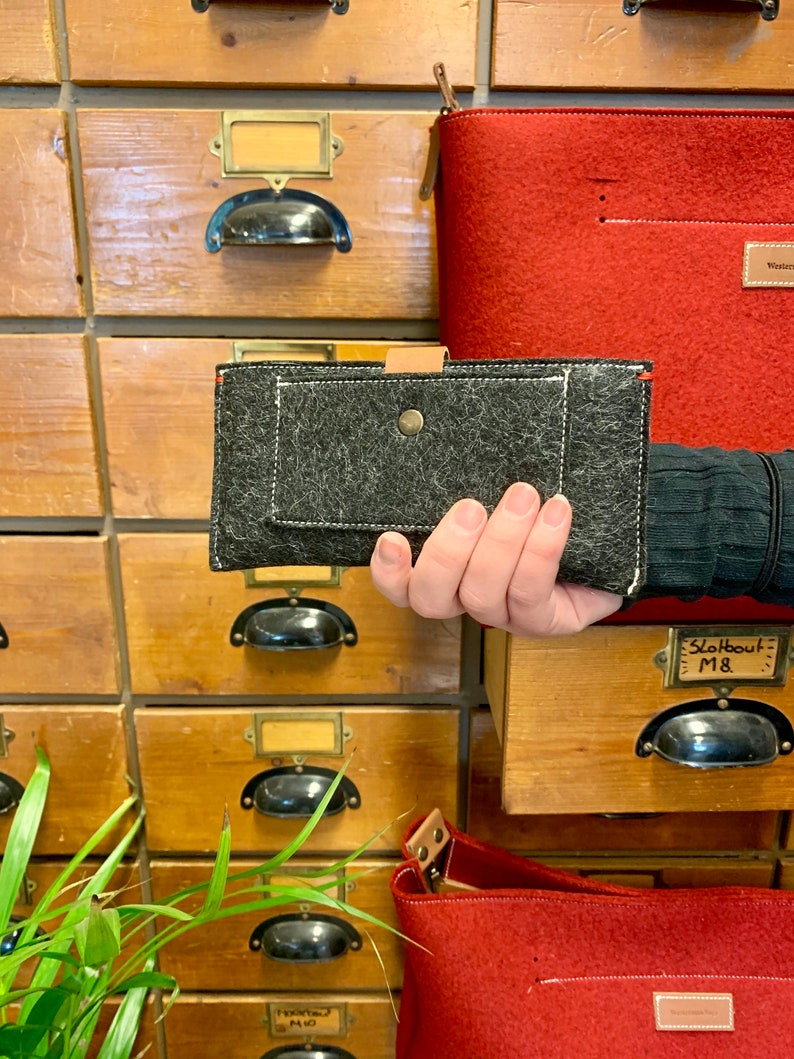 May include: A black felt wallet with a brown leather flap and a white stitch detail. The wallet is held in a hand against a background of wooden drawers.