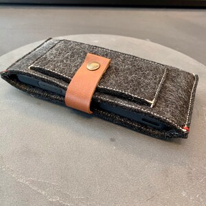 May include: A black felt phone case with a brown leather strap and a metal button closure. The case has a white stitched seam around the edges.