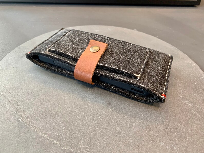 May include: A black felt phone case with a brown leather strap and a metal button closure. The case has a white stitched seam around the edges.
