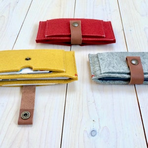 May include: Three felt phone cases in different colors: yellow, red, and gray. Each case has a brown leather strap with a button closure.