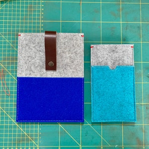 May include: Two felt pouches, one blue and one turquoise, with a grey felt top. The larger pouch has a brown leather strap with a metal buckle. The pouches are on a green cutting mat.