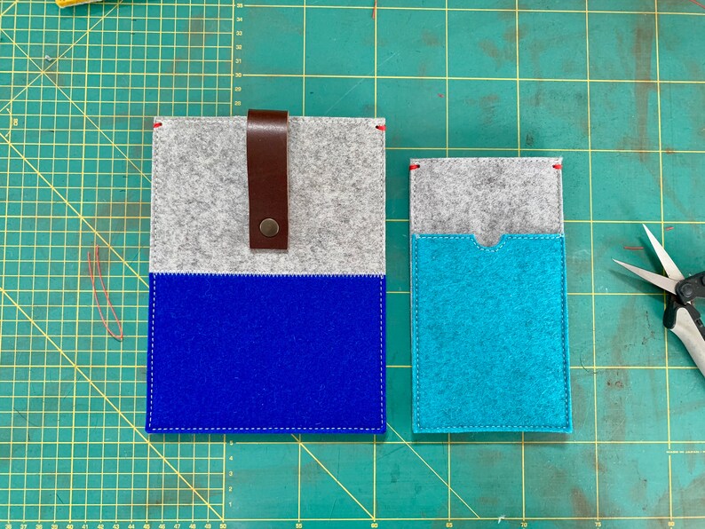 May include: Two felt pouches, one blue and one turquoise, with a grey felt top. The larger pouch has a brown leather strap with a metal buckle. The pouches are on a green cutting mat.