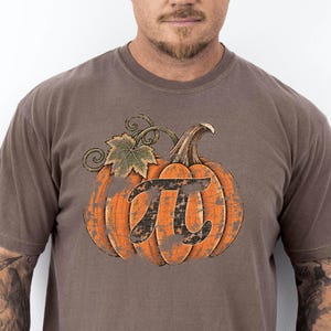 Peut inclure: Un t-shirt marron à manches courtes arborant un grand motif de citrouille orange avec une feuille et une tige vertes. Le centre de la citrouille affiche un symbole mathématique pi noir avec un aspect texturé et usé sur tout le motif.