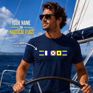 Custom Nautical Flag Shirt in navy blue featuring colorful maritime signal flags across the chest. A man in sunglasses steers a sailboat under a bright blue sky.