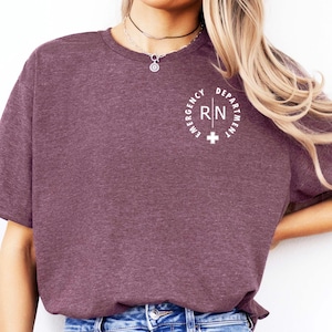 Peut inclure: T-shirt bordeaux à col rond et manches courtes. Le t-shirt présente un motif circulaire blanc sur la poitrine avec l'inscription "EMERGENCY DEPARTMENT RN" et une croix. Le modèle porte un jean bleu.