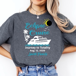 Peut inclure: T-shirt gris foncé avec un motif turquoise et blanc. Le motif comprend les mots "Eclipse Cruise" au-dessus d'une illustration de bateau de croisière, de palmiers et le texte "Journey to Totality Aug. 12, 2026 Smith Family."